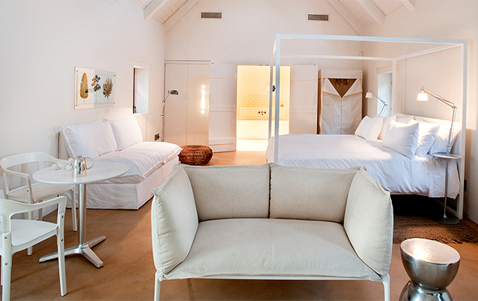 An all-white cottage suite with a slim steel four-poster, a day bed under a framed pressed-plant study, a small round bistro table and chairs and a boxy cream armchair on a polished screed floor, the bathroom lit beyond an open doorway.