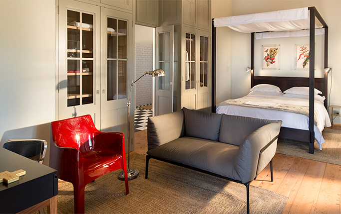 A farmhouse room with a dark timber four-poster under a white canopy, a translucent red moulded chair and a grey sofa on a jute rug, glazed cupboards along one wall and a black and white checkerboard bathroom floor visible through the open door.