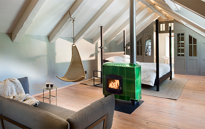 An attic room under whitewashed rafters where a green glazed-tile wood stove burns beside a four-poster bed, a woven hanging chair suspended from the roof beam and a knitted throw over the sofa on wide pale floorboards.