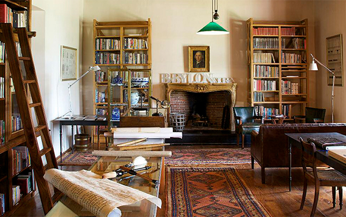 The Manor House library, floor-to-ceiling timber shelves of books served by a rolling ladder, a portrait hung above a marble fireplace with wooden BABYLONSTOREN letters on the mantel, worn Persian rugs and rolled botanical charts across the tables.