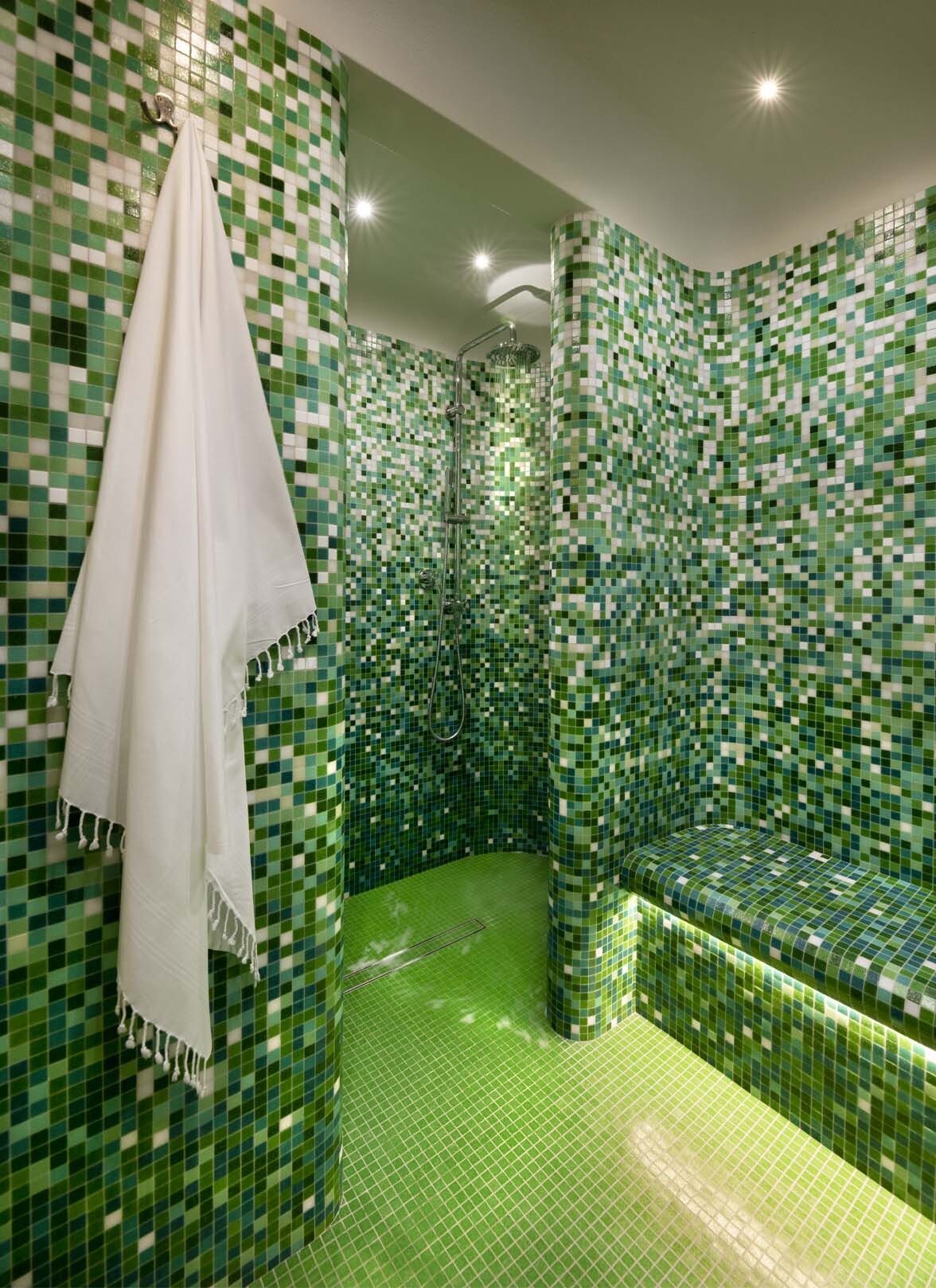 A steam room tiled floor to ceiling in mixed green mosaic, curved partition walls around a rain shower head, a built-in bench lit from below and a white fringed towel hanging by the entrance.